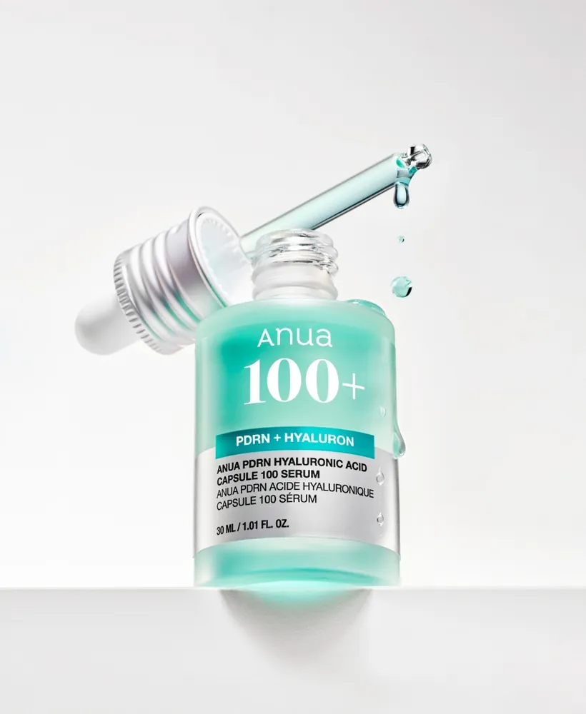 A mint-green Anua '100+' serum bottle with a silver label band reading 'PDRN + Hyaluron', standing on a white ledge with its silver dropper cap lifted out at an angle and drops falling, on a light grey background.