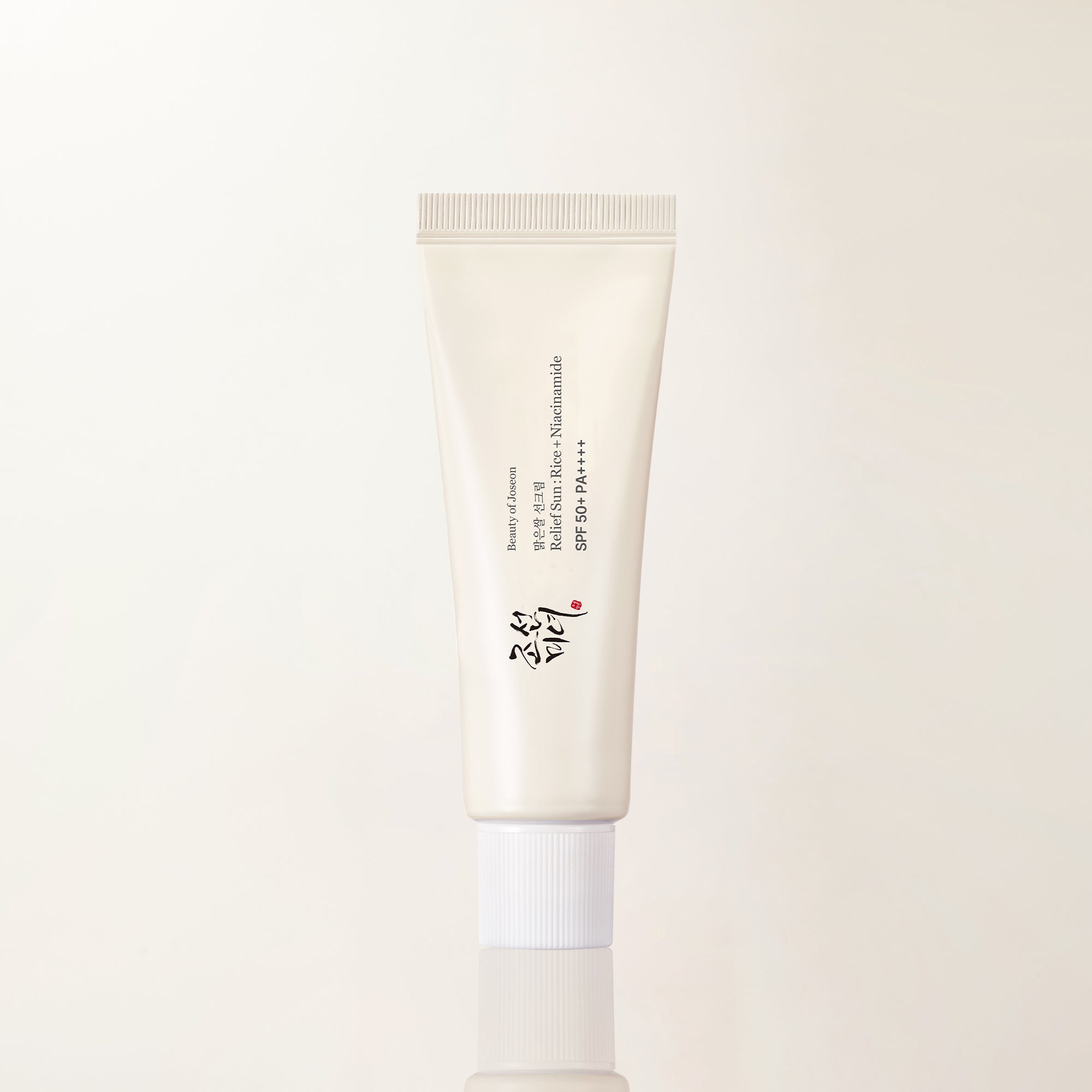 Tubo color crema con tapón blanco estriado, con la etiqueta «Beauty of Joseon, 맑은쌀 선크림, Relief Sun : Rice + Niacinamide, SPF 50+ PA++++» y el logotipo caligráfico de la marca, sobre una superficie beige reflectante.
