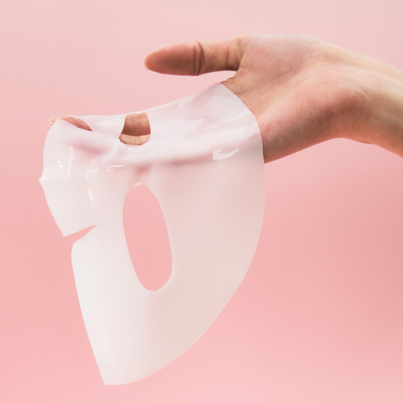Una mano sostiene una mascarilla facial de hidrogel translúcida, rosa lechosa, con aberturas para ojos y nariz, sobre fondo rosa.