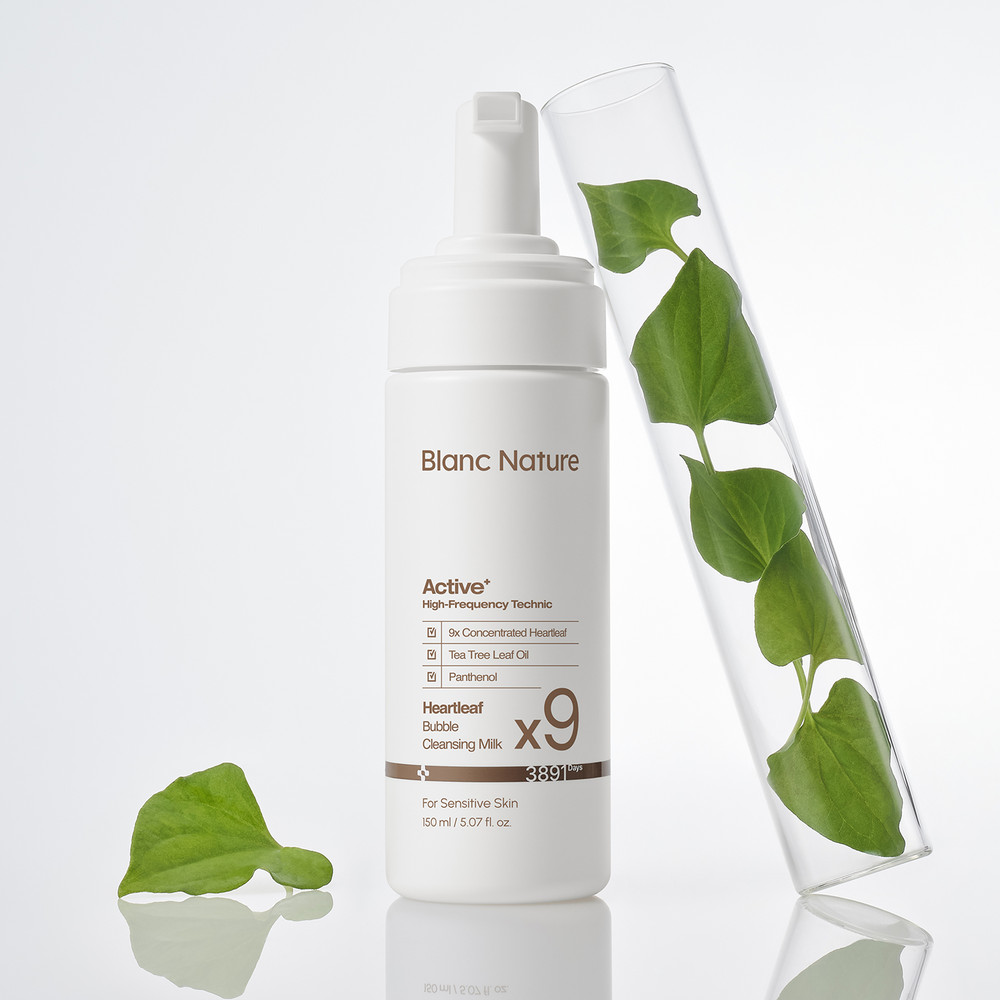 Frasco blanco con dosificador de espuma (texto marrón y un gran «x9») junto a un tubo de vidrio con hojas verdes de heartleaf y una hoja suelta sobre una superficie blanca reflectante.