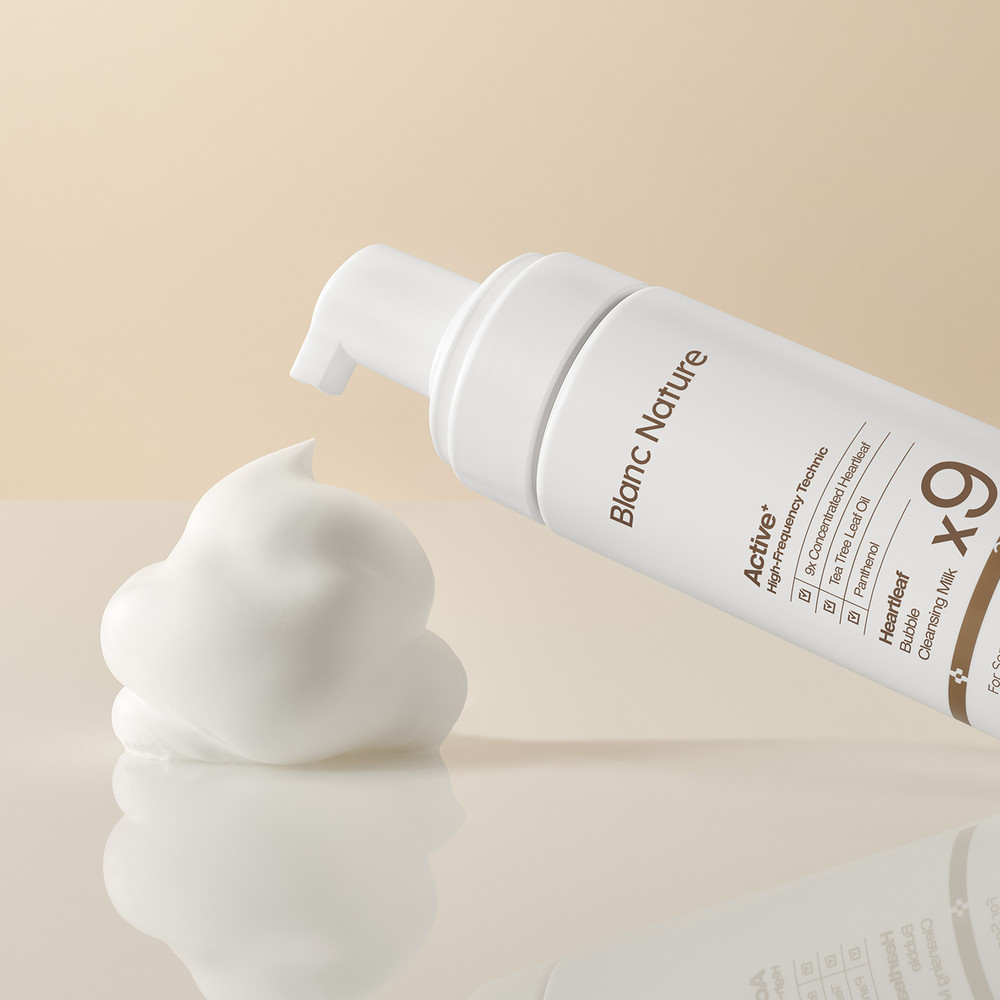 Un copete espeso de espuma blanca sobre una superficie crema brillante, con el dosificador del frasco inclinado hacia él sobre fondo beige.
