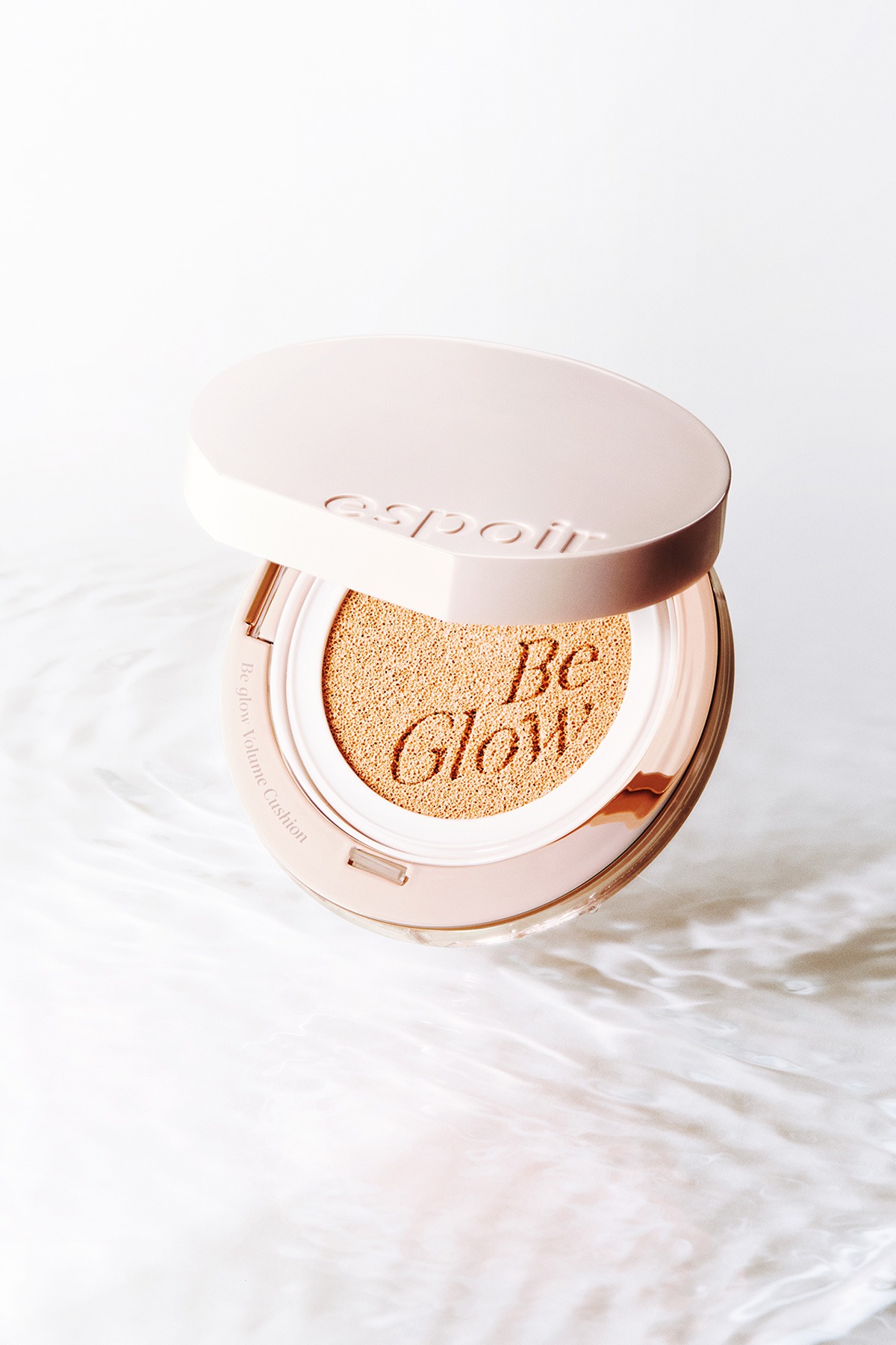 An open pale-pink espoir Be Glow Volume Cushion compact floating above rippled white water, the lid embossed with 'espoir' and the beige cushion embossed with 'Be Glow'.