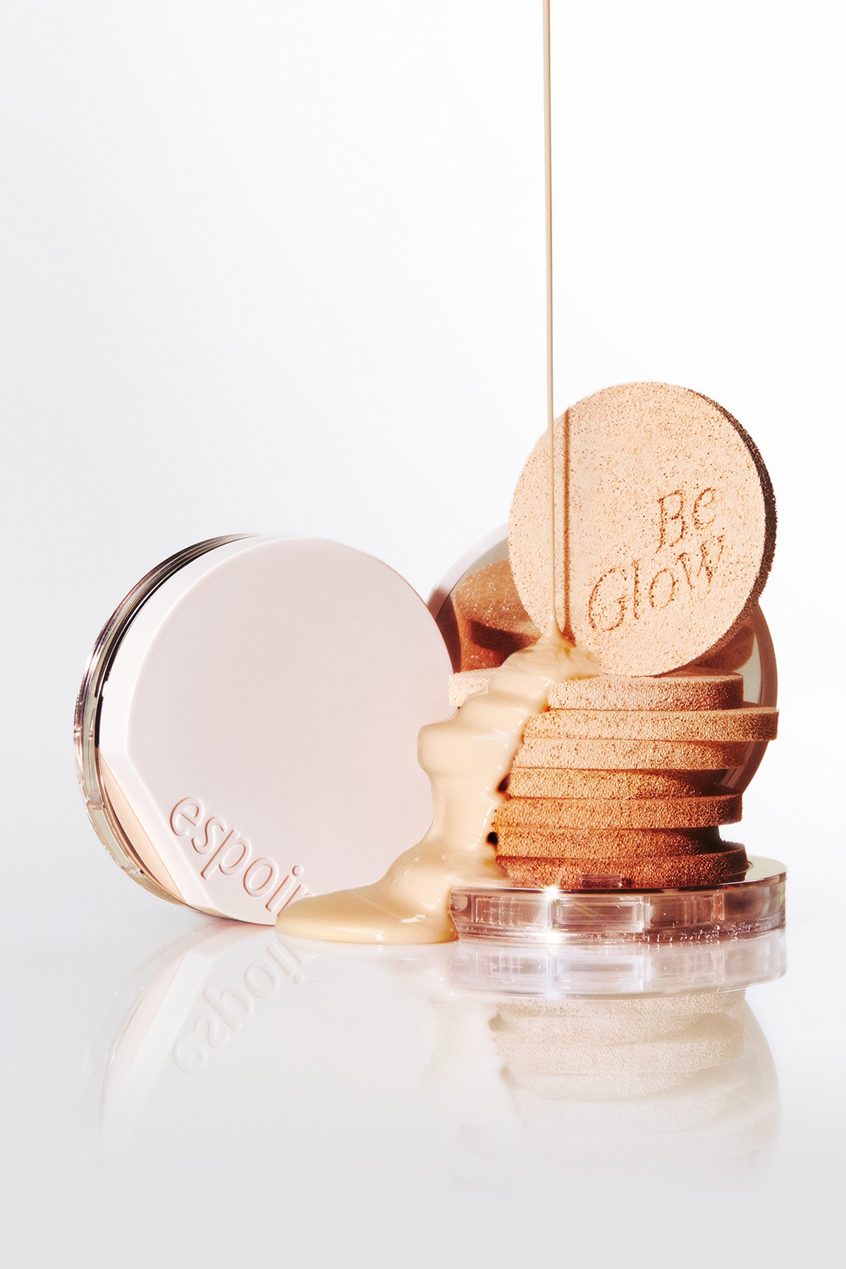 Light beige liquid foundation pouring over a stack of cushion puffs beside a closed pale-pink espoir compact, with one puff embossed 'Be Glow'.