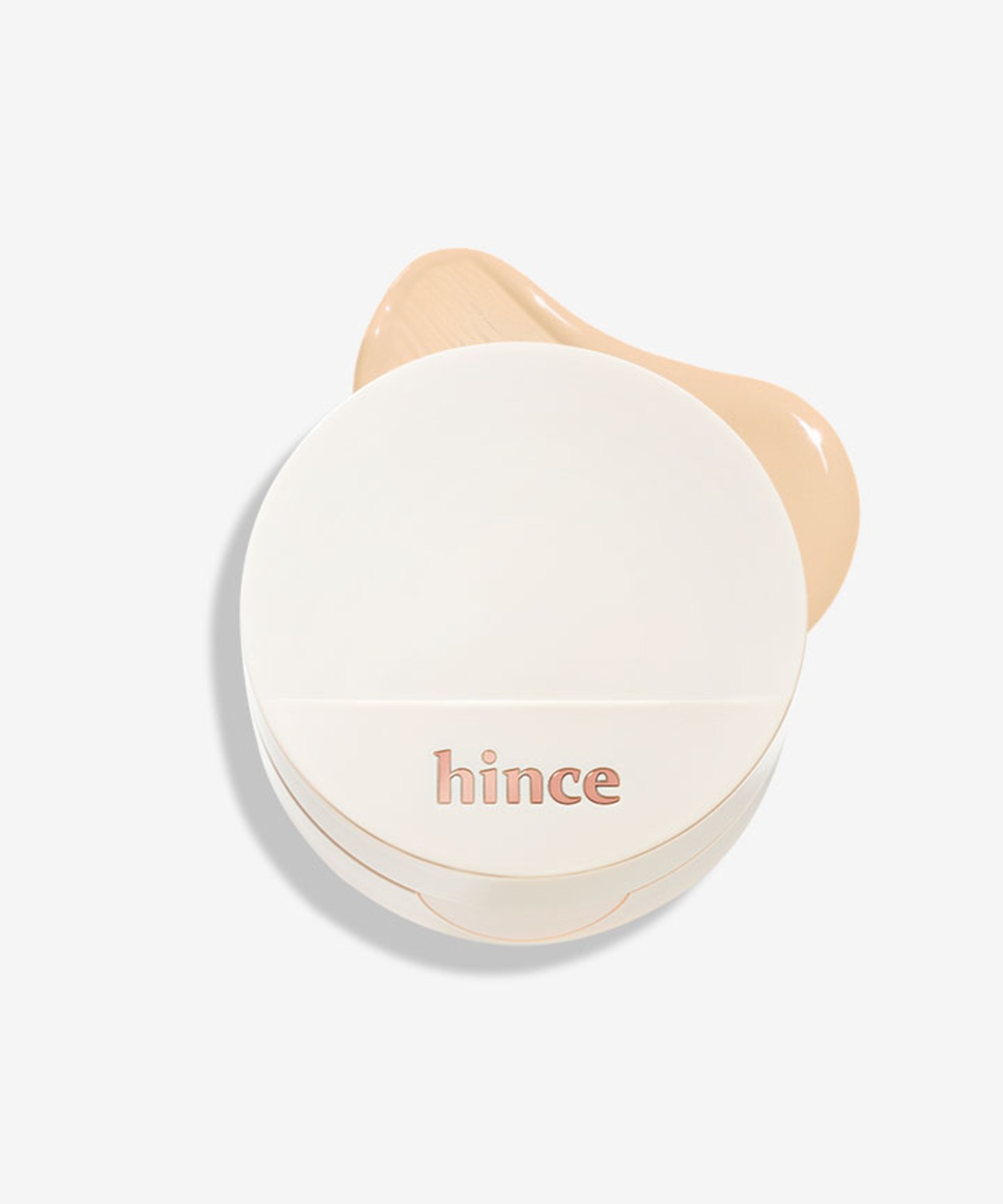 A closed cream-white hince Second Skin Glow Cushion compact with a rose-gold 'hince' logo, in front of a smooth swoosh of beige foundation on a light grey background.