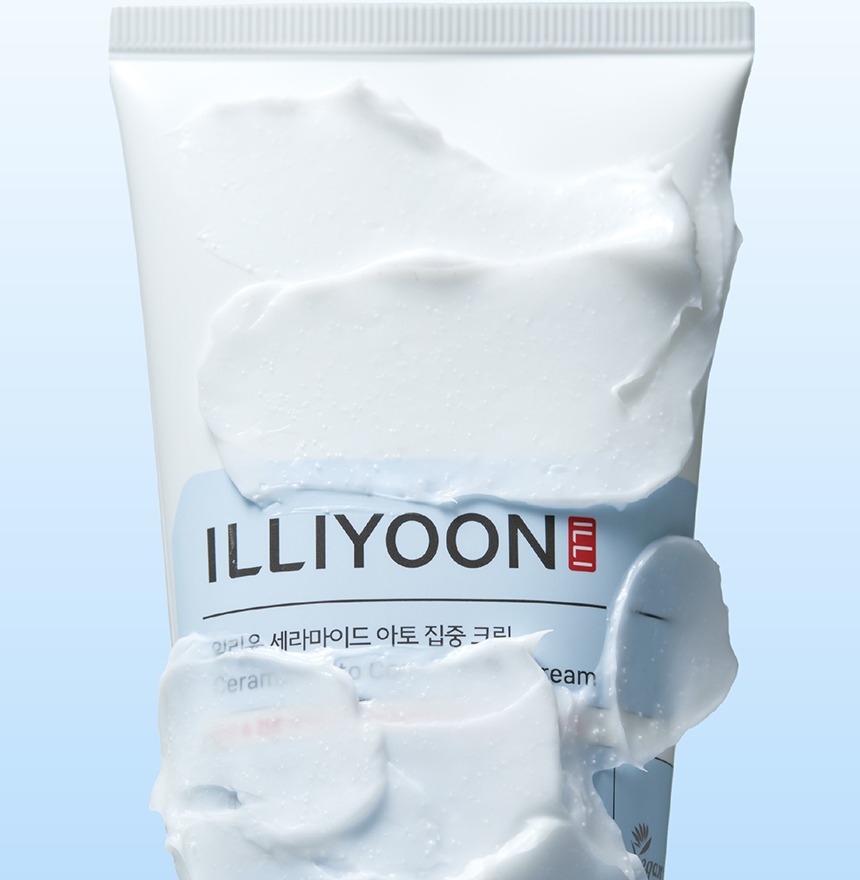 Crema blanca y espesa extendida en amplias pasadas sobre la parte frontal de un tubo blanco de ILLIYOON, cubriendo en parte su etiqueta azul claro, sobre fondo azul pálido.