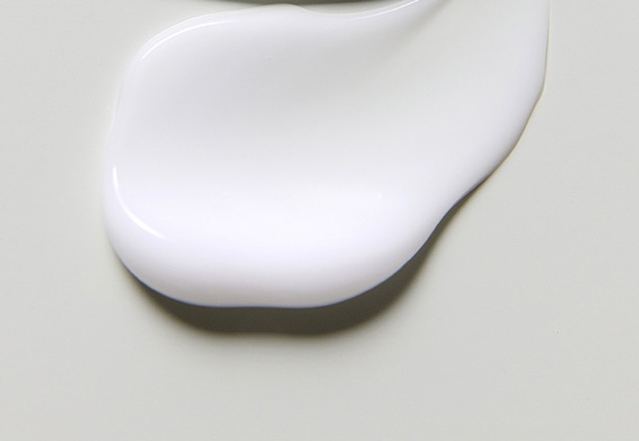 Una porción espesa y brillante de leche limpiadora blanca extendida sobre una superficie gris verdosa pálida.