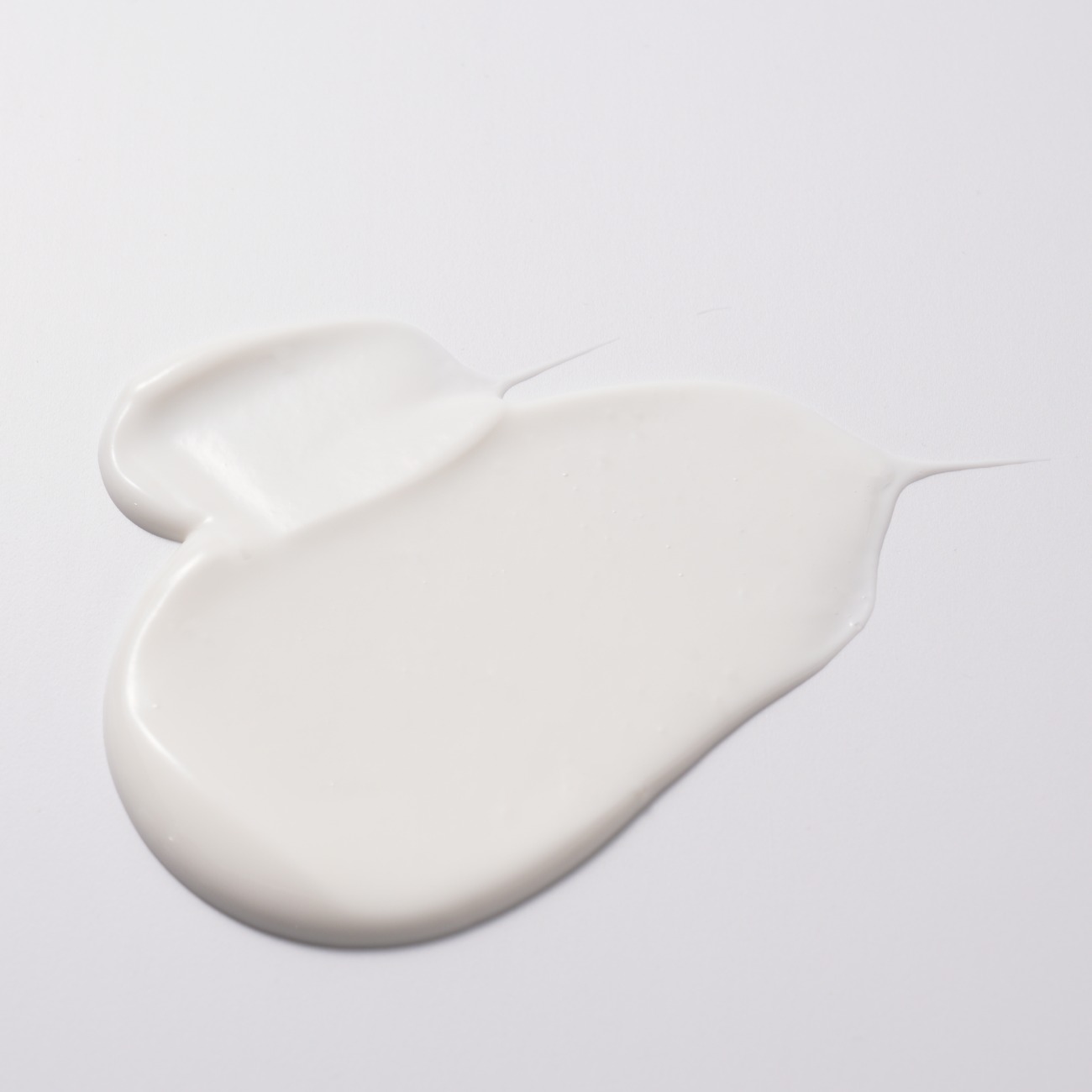 A thick, glossy swirl of white cleansing milk spread on a light-grey surface, with two thin trailing wisps at the edges.