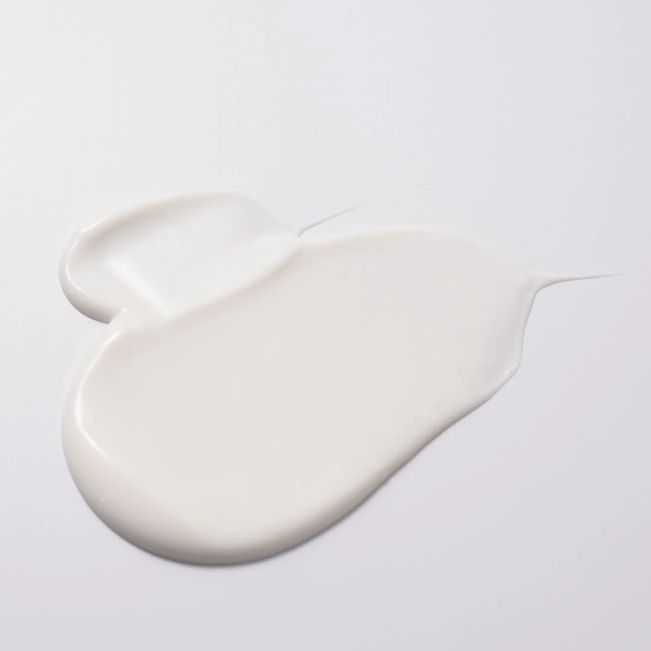 Una mancha espesa y brillante de leche limpiadora blanca extendida sobre una superficie gris claro, con dos hilos finos en los bordes.