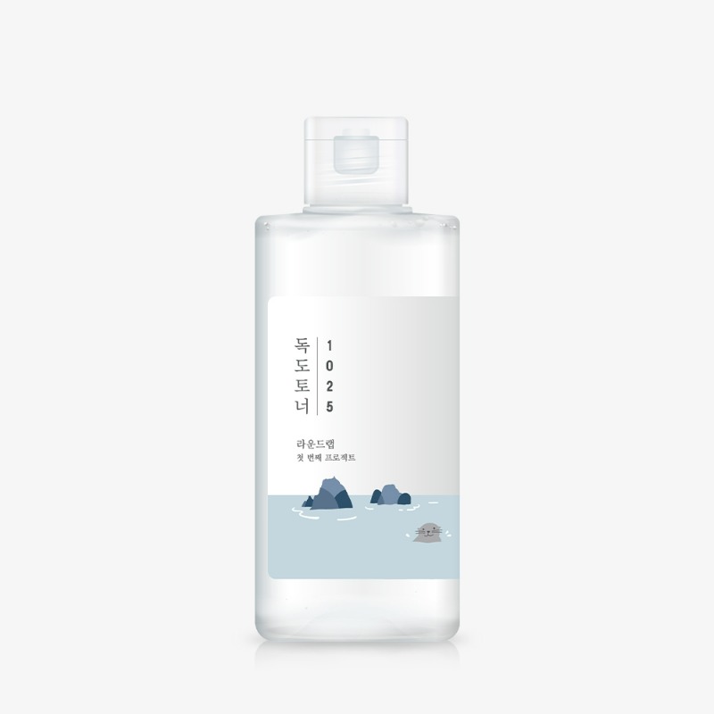 Botella de plástico transparente con tapa abatible esmerilada y etiqueta blanca con el texto vertical «독도토너 1025» y una pequeña ilustración de islotes rocosos y una foca, sobre fondo gris claro.