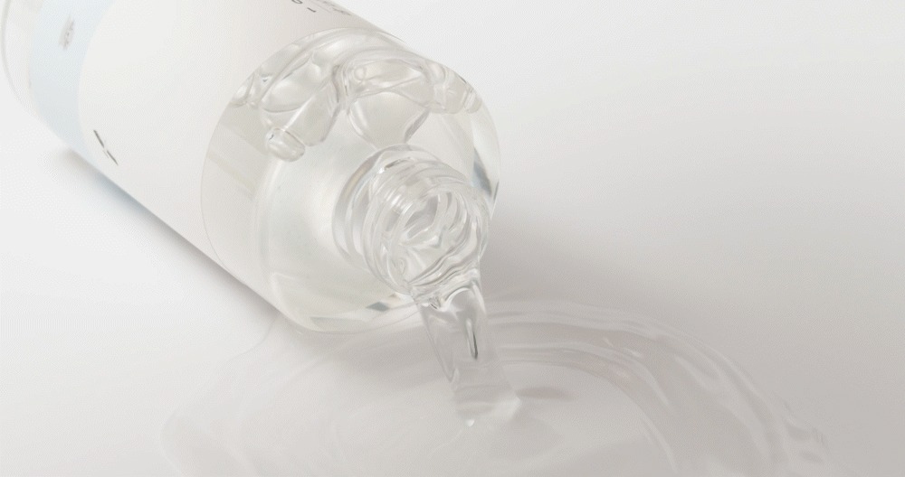Una botella transparente tumbada y sin tapón de la que sale un líquido transparente y acuoso que se extiende en un charco fino y brillante sobre una superficie blanca.