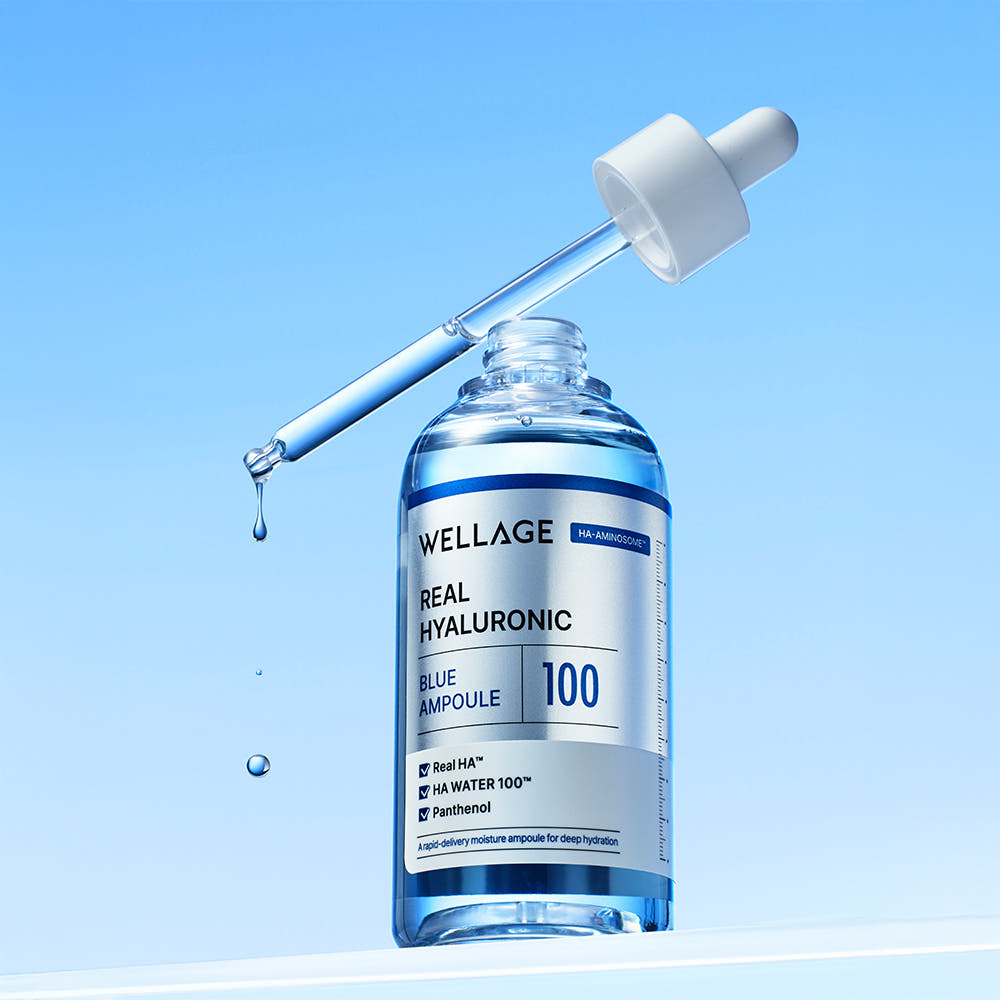 Frasco azul transparente de WELLAGE Real Hyaluronic Blue Ampoule 100 con etiqueta plateada y el cuentagotas levantado sobre el cuello del que caen gotas, sobre fondo azul claro.